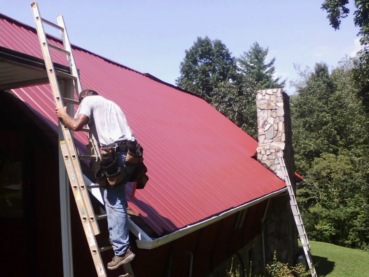 Roofer on a ladder beside a red metal roof and stone chimney — Soffit & Fascia Repair by Roofing Brothers in East Sandwich